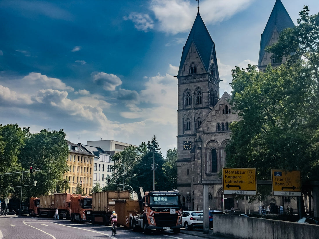 A cyclist riding past orange dump trucks near a stone church in Koblenz